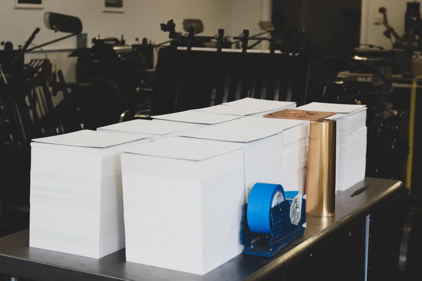 Stacks of cut cotton paper blanks on the press-room table beside a roll of metallic foil and a copper die, with letterpress presses in the background